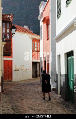 Une femme âgée vêtue de noir marchant dans une rue pavée aux façades colorées et un balcon en bois à Agulo, la Gomera, îles Canaries, Espagne Banque D'Images