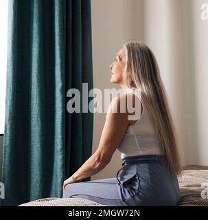 Vue latérale d'une femme triste et âgée assise sur un lit à la maison et regardant la fenêtre. Femme solitaire pensive senior dans la chambre. Banque D'Images