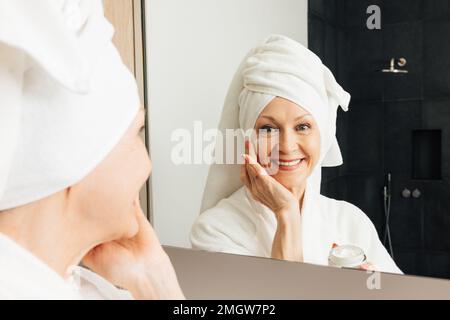 Portrait d'une femme aînée souriante appliquant un traitement du visage. Femme d'âge gaie avec crème faciale dans la salle de bains. Banque D'Images