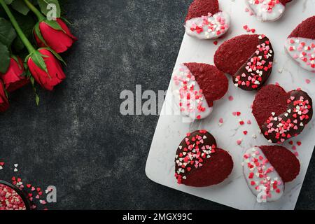 Des biscuits au velours rouge ou au brownie sur le cœur en forme de glaçage au chocolat avec des roses rouges sur fond noir. Idée de dessert pour la Saint-Valentin, les mères ou les femmes Banque D'Images