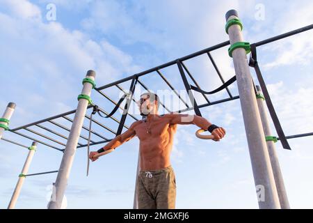 Un athlète s'entraîne avec des anneaux de gymnaste dans une salle de sport extérieure, homme en excellente forme physique Banque D'Images