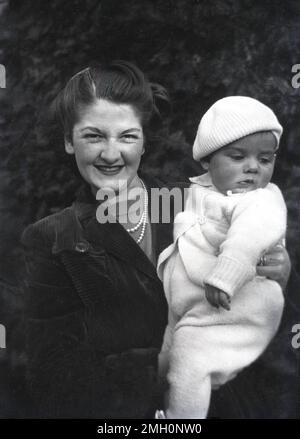 Fin 1940s, historique, une mère portant une veste en velours côtelé et un collier de perles debout à l'extérieur tenant son enfant en bas âge, qui est dans un body et un chapeau de béret, Angleterre, Royaume-Uni. Banque D'Images