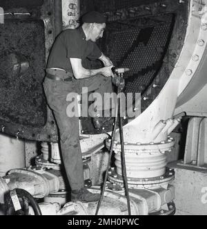 1950s, historique, un ouvrier de l'acier portant un chapeau de béret, au travail, utilisant un pistolet électronique pour introduire des trous de goujon dans des machines géantes circulaires en fer. Abbey Works, Port Talbot, pays de Galles du Sud, Royaume-Uni. Banque D'Images