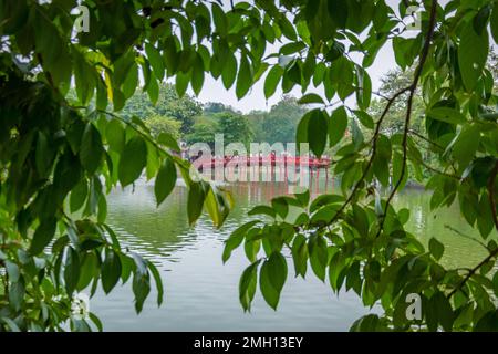 Pont rouge au-dessus d'un lac d'étang dans le parc à Hanoi encadré par des feuilles vertes et des branches d'arbre, Vietnam. Banque D'Images