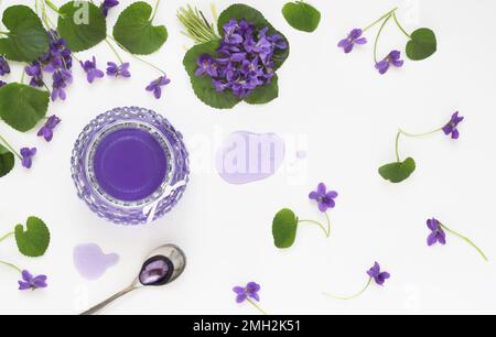 Sirop et gelée de viola odorata fleurs en verre, cuillère et violet fleur fraîche avec feuilles décoratives disposées sur table blanche. Gelée de violette aux herbes Banque D'Images