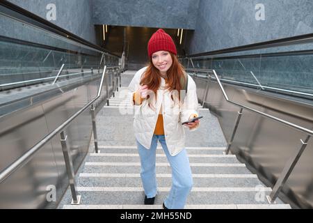 Portrait redhead fille touriste, monte les escaliers avec smartphone, suit l'itinéraire sur l'application de téléphone mobile, tient sac à dos et sourit Banque D'Images