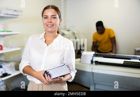 Portrait d'une employée amicale de la maison d'impression avec pile de carnets dans ses mains Banque D'Images