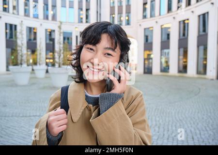 Portrait d'une fille asiatique souriante parle sur téléphone mobile, parle avec un ami sur smartphone, marche dans le centre-ville, se tient dans la rue et rit Banque D'Images