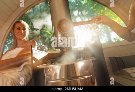 Une femme heureuse se détendant et versant de l'eau sur des pierres chaudes dans un sauna à baril en bois. Vapeur sur les pierres, spa et concept de bien-être, les amies se détendent Banque D'Images