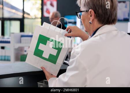 Un employé de pharmacie senior se tenant au comptoir de la pharmacie et plaçant le traitement d'ordonnance dans un sac en papier préparant le colis pour le client. Les clients regardant les étagères pleines de médicaments, achetant des pilules Banque D'Images