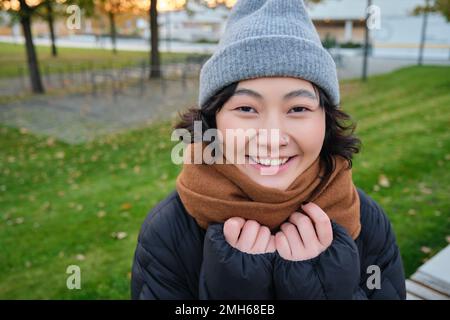 Portrait d'une adorable fille asiatique en chapeau et foulard, promenades en ville par temps froid, sourires et air heureux, assis dans le parc près de l'herbe verte Banque D'Images