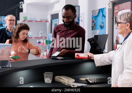 Client afro-américain mettant le téléphone sur le terminal de pos payer pour des suppléments en pharmacie après avoir discuté de soins de santé avec le pharmacien. Client de pharmacie acheter des pilules de prescription Banque D'Images