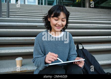 Fille souriante, graphiste, utilisant la tablette numérique et l'outil de stylo pour dessiner, fait l'affectation à domicile pour l'université, assis sur les escaliers Banque D'Images