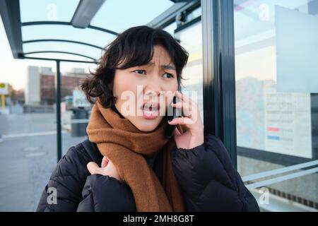 La jeune femme semble frustrée et déçue, parle sur téléphone portable, se tient sur l'arrêt de bus, reçoit de mauvaises nouvelles Banque D'Images