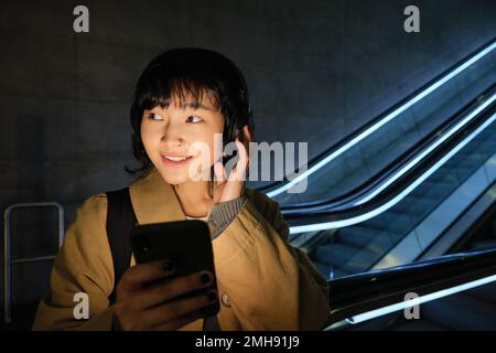 Portrait de la belle fille coréenne dans un casque, se tient près de l'escalier roulant, voyage, se rend à la maison dans les transports en commun, tient le smartphone, pose avec Banque D'Images