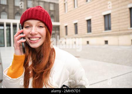 Élégante et moderne, cette jeune fille à tête rouge parle sur un téléphone portable, passe un appel téléphonique, appelle quelqu'un de l'extérieur sur l'application pour smartphone, se tient dans la rue et sourit Banque D'Images