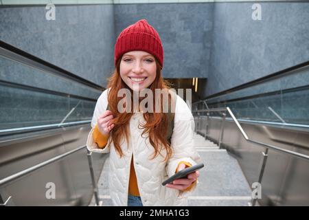 Photo en extérieur d'une jeune femme qui planifie un itinéraire, suit la carte sur l'application pour smartphone, monte les escaliers avec un sac à dos et sourit Banque D'Images