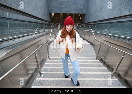 Photo en extérieur d'une jeune femme qui planifie un itinéraire, suit la carte sur l'application pour smartphone, monte les escaliers avec un sac à dos et sourit Banque D'Images