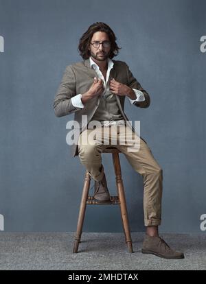 Le style est mon deuxième prénom. Portrait en studio complet d'un jeune homme beau et élégant, assis sur un tabouret sur un fond gris. Banque D'Images