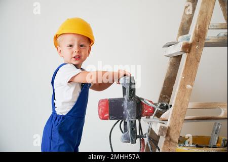 Adorable petit ouvrier de construction de garçon tenant la perceuse électrique et regardant la caméra. Enfant mignon portant une combinaison de travail et un casque de sécurité tout en se tenant sur une échelle contre un mur blanc. Banque D'Images