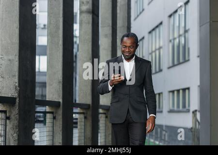 Un homme afro-américain mature et réussi en costume d'affaires marche à l'extérieur d'un immeuble de bureaux, en utilisant un téléphone, en tapant un message et en regardant les nouvelles en ligne tout en souriant Banque D'Images