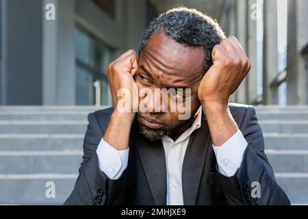 Contrarié afro-américain homme assis sur les escaliers à l'extérieur de l'immeuble de bureaux, homme d'affaires en faillite dans le désespoir, patron mature déprimé dans un costume d'affaires à l'extérieur. Banque D'Images