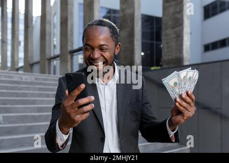 Afro-américain homme en costume d'affaires se réjouissent, a obtenu en ligne prêt de petite entreprise, propriétaire avec de l'argent liquide dollars heureux avec le résultat de réalisation, patron tenant de l'argent comptant et téléphone. Banque D'Images