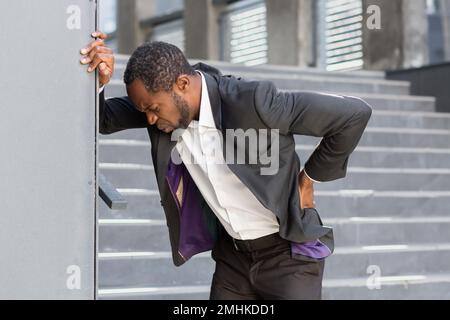 L'homme afro-américain surchargé et malade à l'extérieur du bâtiment de bureau a de graves maux de dos, homme d'affaires massant son côté, patron mature dans les airs près du bureau. Banque D'Images