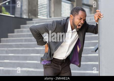 L'homme afro-américain surchargé et malade à l'extérieur du bâtiment de bureau a de graves maux de dos, homme d'affaires massant son côté, patron mature dans les airs près du bureau. Banque D'Images
