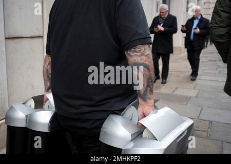 Un ouvrier avec des avant-bras tatoués porte des poubelles de bureau devant des travailleurs adaptés dans la City de Londres, le quartier financier de la capitale, le 26th janvier 2023, à Londres, en Angleterre. Banque D'Images