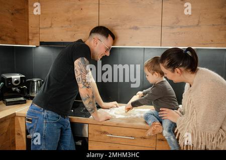 Parents heureux avec bébé fils ensemble sortir la pâte crue avec la goupille sur le plan d'examen de cuisine, vue latérale. Cuisine familiale. Banque D'Images