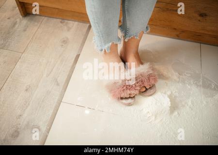 Farine dispersée sur le sol de la cuisine. Pieds femelles en pantoufles sales dans la farine blanche pour la cuisson. Chaussures confortables pour la maison. Banque D'Images