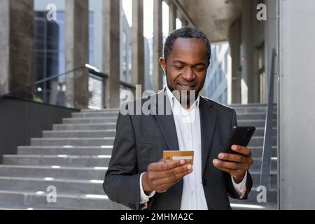 Homme afro-américain heureux et réussi en costume d'affaires à l'extérieur de bâtiment de bureau tenant la carte de crédit de banque et le téléphone, homme d'affaires shopping en ligne et de transférer de l'argent à l'aide de l'application. Banque D'Images