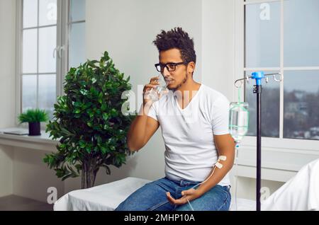 Jeune indien assis dans la salle de médecine sous le compte-gouttes et en buvant de l'eau. Banque D'Images