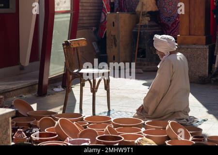Vieux arabe méconnaissable avec une tenue traditionnelle, priant et regardant le sol tout en vendant des bols en argile faits main dans le marché de rue Banque D'Images