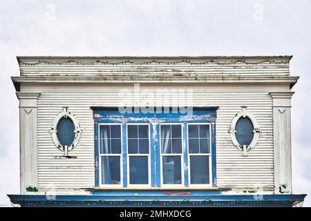 Gros plan sur le deuxième étage d'une vieille belle banque inutilisée avec grande fenêtre carrée entre deux fenêtres ovales dans un cadre en bois patiné Banque D'Images