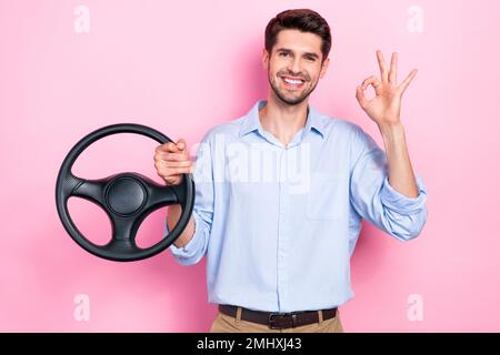 Photo du jeune homme de cheveux brunet porter une chemise de bureau bleue montrer le signe clé de main tenir le volant après avoir obtenu le permis de conduire isolé sur fond rose Banque D'Images