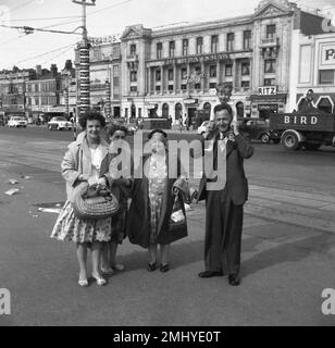 Années 1950, historique, une famille ouvrière debout pour une photo sur la promenade de Blackpool, Angleterre, Royaume-Uni. Dans la rue derrière, sur la Royal Princes Parade, les voitures de l'époque. Le Huntsman, Ritz, Tassauds (Waxworks) sont des noms sur les bâtiments. La famille est habillée formellement, comme les gens sont en vacances sur cette époque, le monsieur, avec un petit garçon sur ses épaules, porte un sit & cravate en trois pièces, et les femmes en robes de fleurs et manteaux longs. Banque D'Images