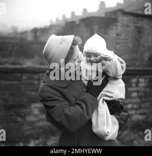 1950, historique, après un baptême, une mère bien habillée dans un manteau de laine, chapeau et gants, avec un grand sourire, à l'extérieur dans un jardin arrière, tenant son bébé heureux, enveloppé dans un body avec chapeau pointu, Kirkheaton, Angleterre, Royaume-Uni, Banque D'Images