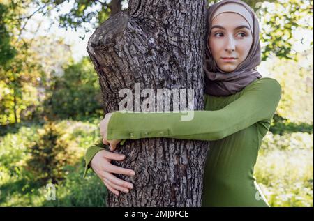 Jeune fille musulmane embrassant un arbre. Gros plan d'une jeune femme dans un hijab et de vêtements décontractés dans le parc. Protéger l'environnement et les soins Banque D'Images