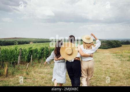 Trois jeunes femmes s'embrasent et marchent sur le vignoble. Les amies blanches vêques et chapeaux ont du plaisir sur le terrain. Bonnes filles ar Banque D'Images