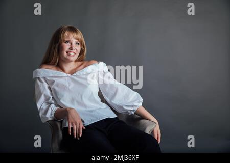 une belle femme dans une chemise blanche est assise sur une chaise avec un fond gris Banque D'Images