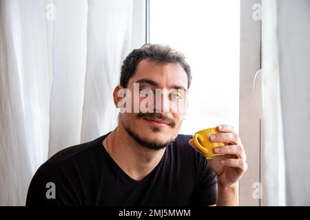 Un jeune homme boit du café assis à la fenêtre. Un bel homme aux yeux bleus tient une tasse jaune et regarde dans l'appareil photo. Banque D'Images