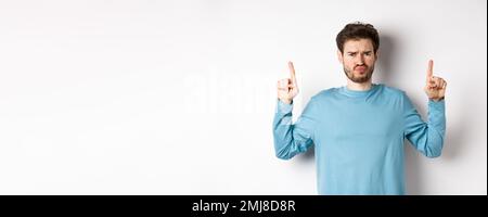 Déçu le modèle masculin frognant et se portant, pointant les doigts vers le haut avec une émotion négative, n'aime pas l'offre promo, se tenant sur fond blanc Banque D'Images