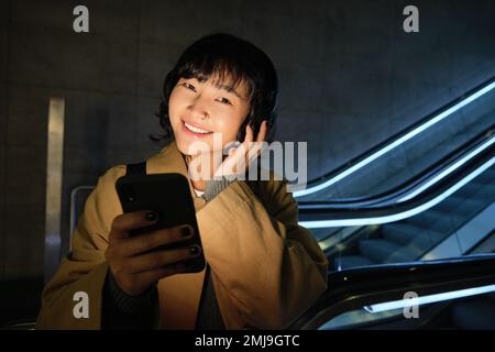 Portrait de la belle fille coréenne dans un casque, se tient près de l'escalier roulant, voyage, se rend à la maison dans les transports en commun, tient le smartphone, pose avec Banque D'Images