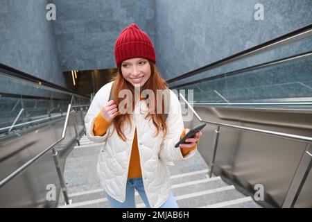 Photo en extérieur d'une jeune femme qui planifie un itinéraire, suit la carte sur l'application pour smartphone, monte les escaliers avec un sac à dos et sourit Banque D'Images