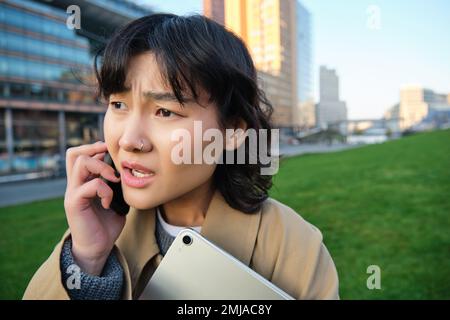 Gros plan sur un portrait d'une jeune femme asiatique qui parle sur un téléphone portable avec un visage inquiet, qui entend de mauvaises nouvelles par conversation téléphonique, qui part de l'université Banque D'Images