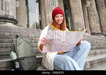 Portrait d'une jeune femme à tête rouge, le touriste se trouve avec une carte papier et cherche un itinéraire vers l'attraction touristique, repose sur des escaliers à l'extérieur Banque D'Images