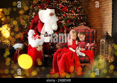 Le Père Noël s'enfile avec des cadeaux tandis que les enfants lisent un livre près de l'arbre de Noël à l'intérieur Banque D'Images