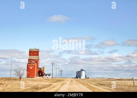 Une route de gravier coupant entre un ancien élévateur de grain en bois rouge et des bacs à grain en acier dans un paysage rural de printemps nuageux Banque D'Images